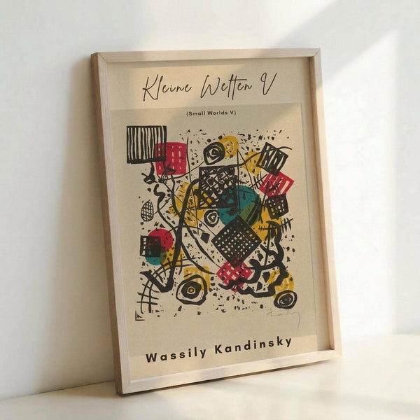 May include: Framed abstract art print in natural wood frame leaning against white wall. Colorful geometric composition with black lines, red squares, yellow shapes, teal circles, and various patterns including stripes and crosshatching. Artist signature "Wassily Kandinsky" visible at bottom. Modern minimalist display setting with soft lighting creating subtle shadows.