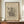 May include: Framed abstract art print in warm brown wooden frame displaying geometric black line drawings and shapes on beige background, positioned on wooden sideboard next to white ceramic vase and stacked books against cream wainscoted wall in modern interior setting.