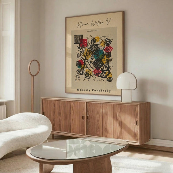 May include: Abstract modernist art print with colorful geometric shapes, patterns, and forms in red, yellow, green, and black displayed in thin wooden frame above walnut credenza. Modern living room scene with white curved chair, glass oval coffee table, and minimalist white table lamp. Warm natural lighting on light colored walls.