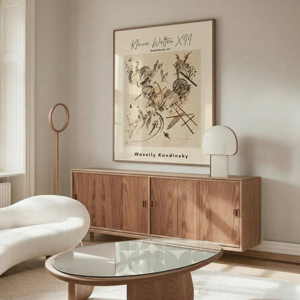 May include: Abstract modernist art print in warm beige tones with dark linear figures and organic shapes, displayed in thin wooden frame above mid-century walnut credenza. Room features cream walls, white upholstered chair, glass-top coffee table, and modern brass floor lamp with spherical shade.