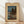 May include: Framed Japanese woodblock print showing a moonlit lake scene with mountains, trees, and traditional buildings reflected in turquoise water. The artwork features a full moon in a dusky blue sky with clouds, dark silhouetted trees, and small figures on a path. Displayed in a light wooden frame with cream matting against white walls on light wood flooring with natural shadows.