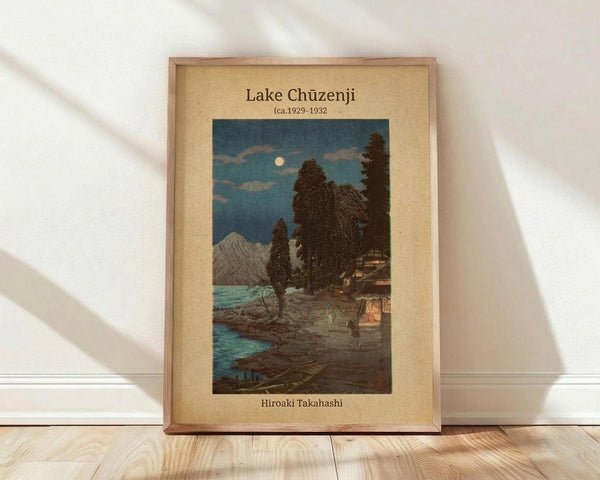 May include: Framed Japanese woodblock print showing a moonlit lake scene with mountains, trees, and traditional buildings reflected in turquoise water. The artwork features a full moon in a dusky blue sky with clouds, dark silhouetted trees, and small figures on a path. Displayed in a light wooden frame with cream matting against white walls on light wood flooring with natural shadows.