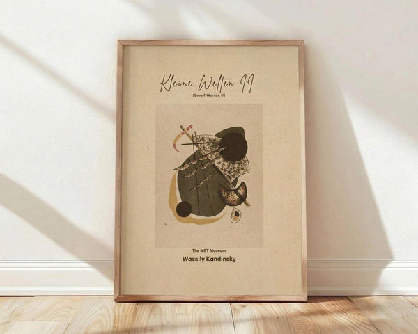 May include: Framed art print leaning against white wall on light wood floor. Cream-colored poster features abstract composition with dark green and black organic shapes, yellow accents, and geometric elements including crosses and circles. Natural wood frame, soft shadows from window light visible on wall and floor.