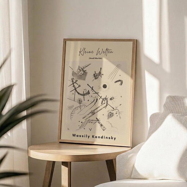 May include: Framed abstract art print with geometric black line drawings on cream background, displayed on round wooden side table next to beige upholstered chair. Natural lighting creates soft shadows across neutral-toned room with plant visible in corner. Artwork features angular shapes, dots, and flowing lines in minimalist style within thin wooden frame.