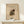 May include: Abstract art poster in wooden frame displaying organic shapes in earthy tones of green, brown, and cream with geometric elements and curved forms. Small wrapped white gift box with natural twine and pine sprig sits beside the framed artwork on light wooden surface against neutral wall.