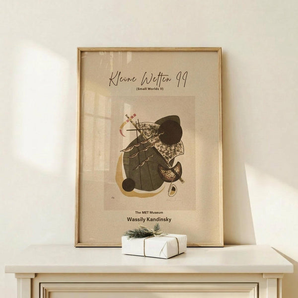 May include: Abstract art poster in wooden frame displaying organic shapes in earthy tones of green, brown, and cream with geometric elements and curved forms. Small wrapped white gift box with natural twine and pine sprig sits beside the framed artwork on light wooden surface against neutral wall.