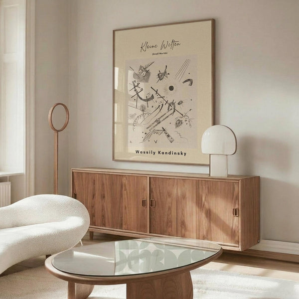 May include: Framed abstract art print with black geometric shapes and lines on cream background, displayed above wooden sideboard in minimalist living room. White curved chair and glass coffee table visible. Warm wood tones throughout modern interior setting.