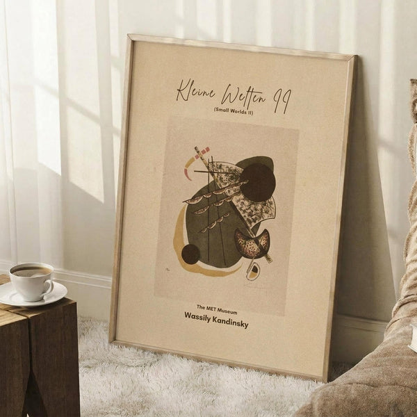 May include: Abstract art print in light wooden frame leaning against white wall corner on white textured rug. Poster features beige background with geometric shapes in olive green, brown, and yellow tones forming an abstract composition. White coffee cup on wooden block visible nearby, cozy blanket draped at edge of scene.