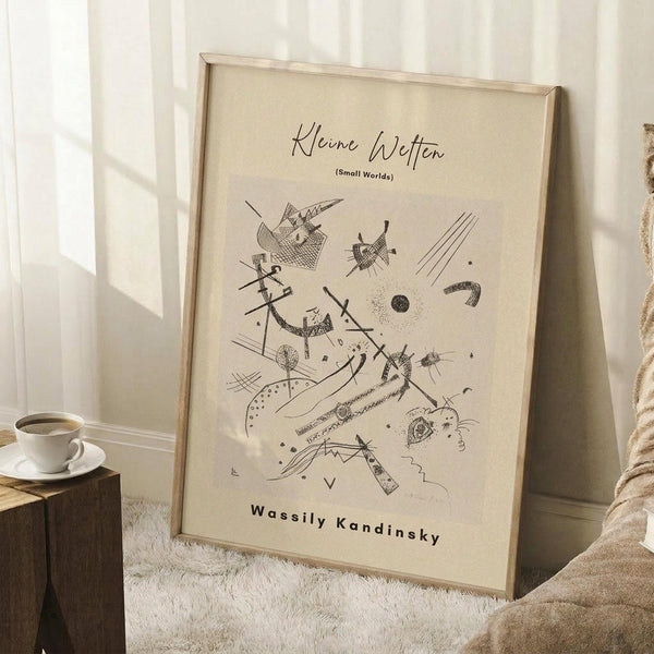 May include: A framed abstract art print with black geometric shapes, lines, and forms on cream paper, displayed in a light wood frame leaning against a white wall. A coffee cup sits on a wooden block beside the artwork, with a textured cream blanket draped nearby on what appears to be a white furry rug.