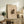 May include: Framed art print with beige background featuring abstract geometric shapes in dark green, yellow, and brown tones, displayed on rustic wooden console table alongside fern plant, books, ceramic vase, and leather armchair in warm neutral living room setting