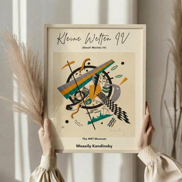 May include: Person holding framed abstract art print featuring geometric shapes, lines and patterns in orange, teal, black and beige colors. Modern Kandinsky-style artwork in light wood frame against white curtained window. Dried pampas grass visible on left side. Person wearing cream colored ruffled sleeve top displays museum poster with angular, dynamic composition.
