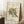 May include: Abstract art poster in light wood frame leaning against white wall corner. Kandinsky-style artwork features geometric shapes, curved lines, and organic forms in teal, orange, black and gray on cream background. Small wooden side table with white coffee cup visible. Textured white rug and beige fabric partially shown. Museum exhibition poster format with text at top and bottom.