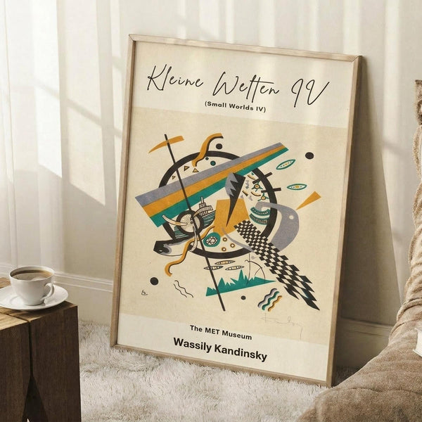 May include: Abstract art poster in light wood frame leaning against white wall corner. Kandinsky-style artwork features geometric shapes, curved lines, and organic forms in teal, orange, black and gray on cream background. Small wooden side table with white coffee cup visible. Textured white rug and beige fabric partially shown. Museum exhibition poster format with text at top and bottom.