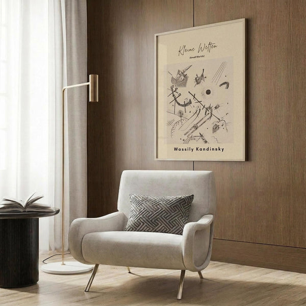 May include: Abstract Kandinsky-style art print in cream and brown tones displayed on wood paneled wall above modern beige armchair with geometric patterned pillow. Scene includes brass floor lamp with white shade, dark round side table with open book, and light wood flooring. Warm, minimalist interior styling with natural lighting from window.