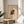 May include: Framed abstract art print featuring organic shapes in black, beige, and mustard yellow with linear details, displayed on a light wood round side table next to a cream upholstered chair. Natural sunlight casts geometric shadows across the neutral-toned room setting with a plant visible in the corner.