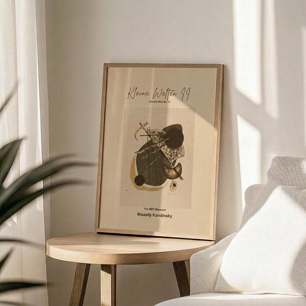 May include: Framed abstract art print featuring organic shapes in black, beige, and mustard yellow with linear details, displayed on a light wood round side table next to a cream upholstered chair. Natural sunlight casts geometric shadows across the neutral-toned room setting with a plant visible in the corner.