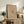 May include: Framed beige abstract art print with geometric line drawings and German text, displayed on rustic wooden sideboard alongside potted fern, ceramic vase, stacked books, and brown leather armchair in neutral-toned living room setting with natural lighting