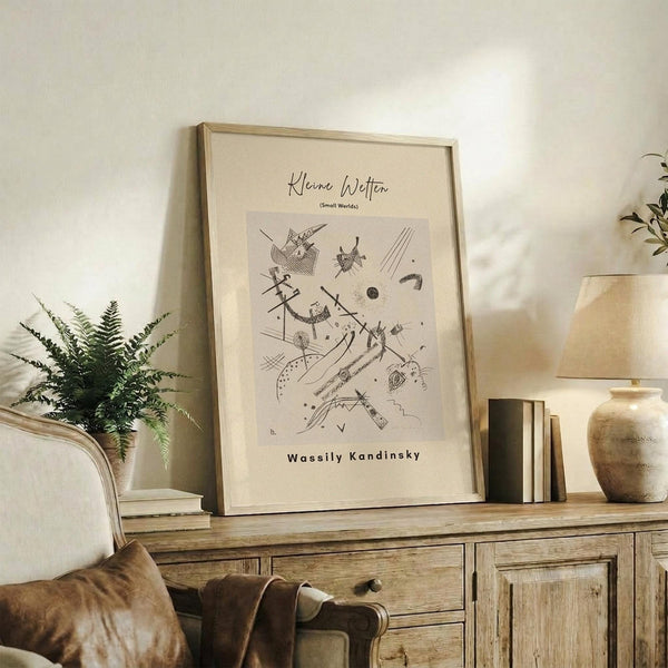 May include: Framed abstract art print with geometric black line drawings on cream background, displayed on rustic wood sideboard with fern plant, leather chair, books, and ceramic vase in warm neutral living room setting