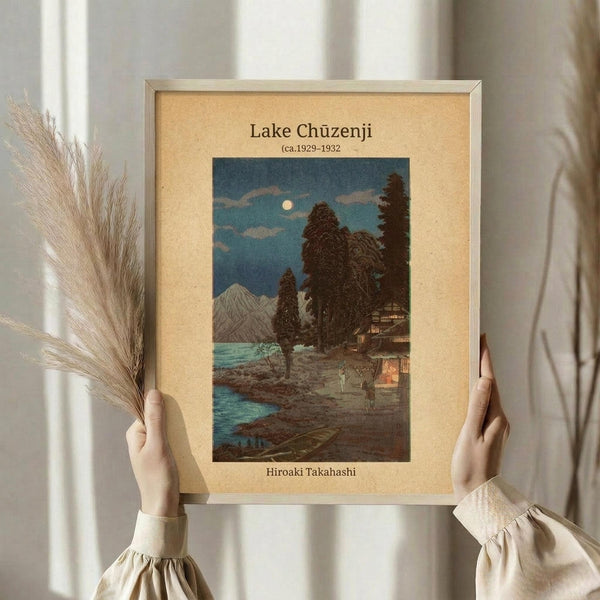 May include: Person in cream-colored ruffled blouse holding framed vintage Japanese woodblock print showing moonlit lake scene with mountains, tall trees, small buildings with warm glowing windows, and turquoise water. Dried pampas grass visible on left side. Print has beige mat border in light wood frame against soft neutral curtained background.