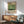 May include: Framed poster with Mark Rothko's name and birth-death dates displayed above color-blocked sections in cream, yellow, and green tones, shown hanging on white wall above wooden sideboard with white ceramic lamp, next to mid-century modern sage green upholstered armchair with wooden frame in sunlit room setting.