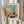 May include: Person in cream sweater holding framed abstract art print with horizontal bands of teal blue, orange, and beige brushstrokes. Light wooden frame against white curtained window background. Dried pampas grass held in other hand. Soft natural lighting creates peaceful, minimalist home decor scene.