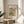 May include: Framed poster with William Morris floral pattern featuring colorful botanical design in blues, greens, coral, and yellow tones displayed on round wooden side table next to beige upholstered chair. Sunlight streams across white wall creating soft shadows from nearby plant leaves in minimalist modern interior setting.