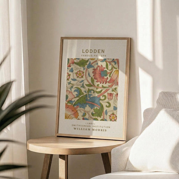 May include: Framed poster with William Morris floral pattern featuring colorful botanical design in blues, greens, coral, and yellow tones displayed on round wooden side table next to beige upholstered chair. Sunlight streams across white wall creating soft shadows from nearby plant leaves in minimalist modern interior setting.