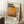 May include: Framed vintage-style poster with Mark Rothko's name in bold black letters above an abstract orange and brown painting. Features biographical text and birth-death years. Light wood frame leaning against white wall next to coffee cup on stack of books and beige textured blanket on white fluffy rug.