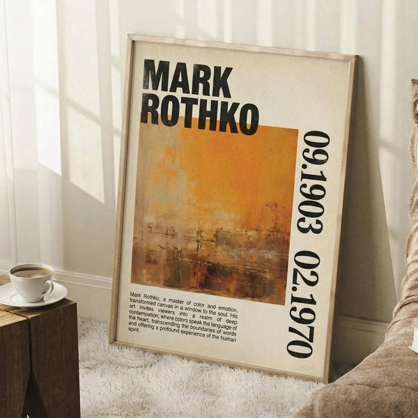 May include: Framed vintage-style poster with Mark Rothko's name in bold black letters above an abstract orange and brown painting. Features biographical text and birth-death years. Light wood frame leaning against white wall next to coffee cup on stack of books and beige textured blanket on white fluffy rug.