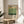 May include: Framed art print with Mark Rothko's name in black text at top, horizontal color blocks in cream, yellow, light green, and dark green, with "Rothko" and dates "09.1903 02.1970" in black text at bottom. Print sits on round wooden side table next to beige upholstered chair, with plant shadow cast on white wall behind. Natural sunlight creates geometric shadows across the scene.