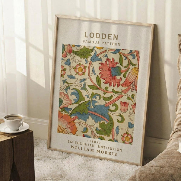 May include: Framed botanical art print with ornate floral pattern in coral, blue, and green tones featuring swirling leaves and decorative flowers. Light wood frame leans against white paneled wall next to wooden side table with white coffee cup. Cream textured rug and beige throw blanket visible in cozy room setting with natural window light casting soft shadows.