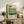May include: Framed vintage-style art poster with Mark Rothko's name and birth-death dates, featuring horizontal color blocks in cream, yellow, and green tones. Displayed on rustic wooden sideboard alongside fern plant, ceramic vase, books, and table lamp in warm, modern living room setting with neutral textiles.