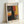 May include: Framed abstract art poster leaning against white wall, featuring bold vertical blocks of orange, red, and yellow paint with black brushstrokes. Light wooden frame contains white matted print with "MARK ROTHKO" text at top and French subtitle. Soft natural lighting creates gentle shadows on textured white wall surface.