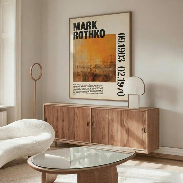 May include: Modern living room with cream walls featuring framed Mark Rothko exhibition poster displaying warm orange and brown abstract painting. Wooden credenza below holds white sculptural lamp, white curved chair and glass oval coffee table in foreground. Minimalist Scandinavian-style interior with natural wood tones and neutral color palette.