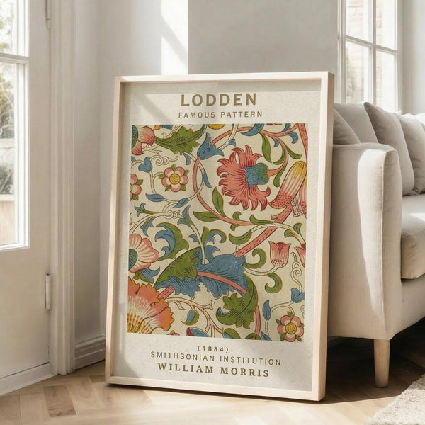 May include: Framed vintage floral pattern print in warm wood frame, leaning against white wall next to beige upholstered sofa. The artwork features intricate botanical design with coral flowers, blue and green leaves, and flowing vines on cream background. Text identifies it as famous Lodden pattern from 1884 by William Morris, Smithsonian Institution. Soft natural lighting streams through tall white-framed windows onto light wooden floors.
