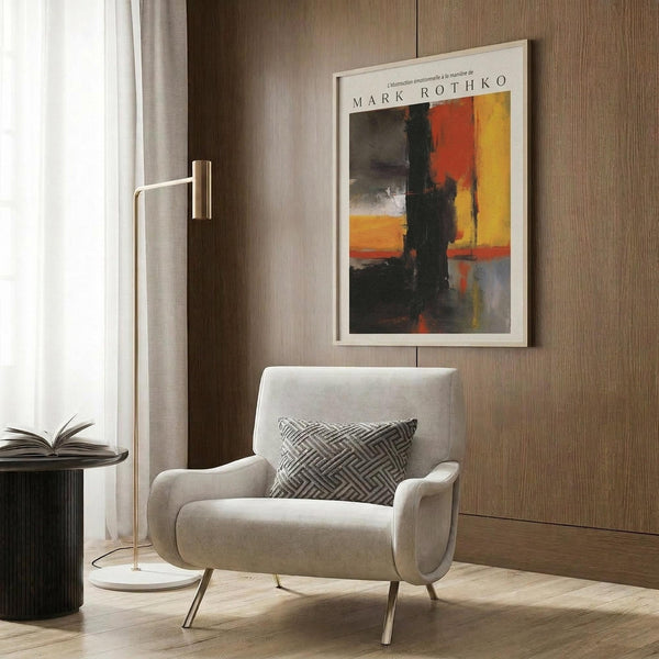May include: Abstract art print with bold orange, red, yellow and black color blocks in white frame, displayed on wood panel wall above modern cream armchair with geometric pillow, brass floor lamp and dark side table visible in contemporary room setting with natural lighting from window