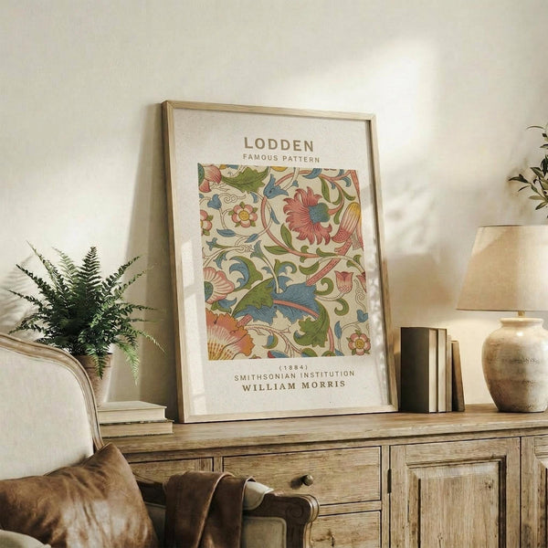 May include: Framed botanical print featuring William Morris's colorful floral pattern with pink flowers, green leaves, and blue accents on cream background. Print displays against white wall on rustic wooden sideboard alongside fern plant, ceramic vase, stacked books, and neutral upholstered furniture in warm, natural lighting.