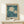 May include: Framed botanical poster with white daisies on teal blue background, displayed on light wood console table against cream wall. Small white wrapped gift with green sprig sits in front of the natural wood frame. Vintage-style art print features hand-drawn flowers and leaves with beige matting and "FLOWER MARKET" and "LISBON" text.