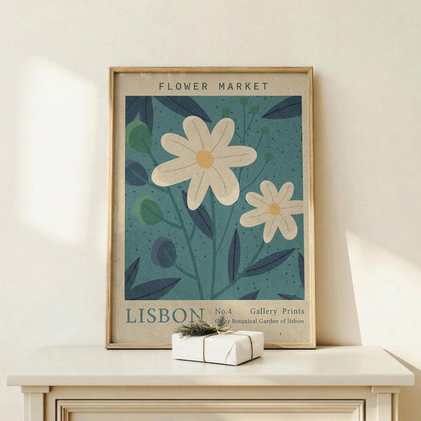 May include: Framed botanical poster with white daisies on teal blue background, displayed on light wood console table against cream wall. Small white wrapped gift with green sprig sits in front of the natural wood frame. Vintage-style art print features hand-drawn flowers and leaves with beige matting and "FLOWER MARKET" and "LISBON" text.