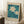 May include: Framed botanical poster with white daisies and blue-green foliage on turquoise background, text reading "Flower Market" and "Lisbon," leaning against white wall corner next to wooden side table with white coffee cup and saucer, cream textured rug beneath