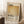 May include: Framed Mark Rothko exhibition poster leaning against white wall corner in sunlit room. Poster features warm beige and golden yellow color blocks with black text showing artist name, dates, and descriptive paragraph. Natural wood frame, positioned on textured white rug next to wooden stool with white coffee cup and saucer. Cozy blanket visible on right side with soft natural lighting creating shadows.