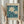 May include: Person in cream ruffled sleeves holding framed botanical poster featuring white daisies on teal background with dark blue leaves, alongside dried wheat stems. Natural wood frame with beige matting displays "FLOWER MARKET" and "LISBON" text. Soft window lighting creates gentle shadows on neutral wall.