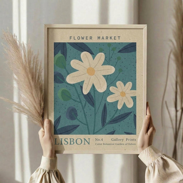 May include: Person in cream ruffled sleeves holding framed botanical poster featuring white daisies on teal background with dark blue leaves, alongside dried wheat stems. Natural wood frame with beige matting displays "FLOWER MARKET" and "LISBON" text. Soft window lighting creates gentle shadows on neutral wall.