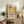 May include: A framed Mark Rothko exhibition poster featuring warm golden and cream abstract color blocks with black typography displaying the artist's name and dates. The print sits on a rustic wooden sideboard alongside a fern plant, ceramic vase, and books in a neutral-toned living room setting with a beige armchair and white walls.