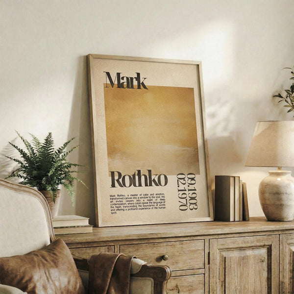 May include: A framed Mark Rothko exhibition poster featuring warm golden and cream abstract color blocks with black typography displaying the artist's name and dates. The print sits on a rustic wooden sideboard alongside a fern plant, ceramic vase, and books in a neutral-toned living room setting with a beige armchair and white walls.