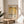 May include: A framed poster with Mark Rothko's name and exhibition details in black text on cream background, featuring golden yellow abstract color field artwork. The wooden-framed print sits on a round wooden side table next to a beige upholstered chair, with plant leaves visible and natural sunlight casting shadows across white walls in a minimalist living room setting.