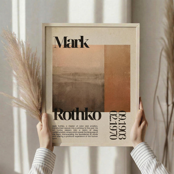 May include: Hands holding a framed beige poster featuring abstract color blocks in warm tones of peach, orange, and brown. The design displays "Mark Rothko" at the top with biographical text and "1903-1970" dates. Natural pampas grass stems visible on the left side, soft window lighting in background creating warm atmosphere in neutral-toned room setting.