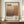 May include: Framed poster with Mark Rothko's name in large black serif letters above abstract geometric blocks in warm brown and orange tones. Features biographical text and birth-death years 1903-1970 in vertical typography. Displayed on wooden sideboard next to white ceramic vase and stacked books against cream walls with white trim molding.