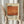 May include: Person holding framed abstract art poster with orange and rust colored rectangular brushstroke painting. Black text reads "Mark Rothko" and includes years "1903-1970" with descriptive paragraph about the artist. Cream colored background with beige wooden frame. Person wears neutral beige clothing and holds dried pampas grass stems. Soft natural lighting against white curtained window background.
