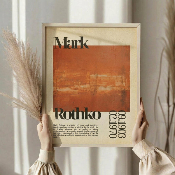 May include: Person holding framed abstract art poster with orange and rust colored rectangular brushstroke painting. Black text reads "Mark Rothko" and includes years "1903-1970" with descriptive paragraph about the artist. Cream colored background with beige wooden frame. Person wears neutral beige clothing and holds dried pampas grass stems. Soft natural lighting against white curtained window background.